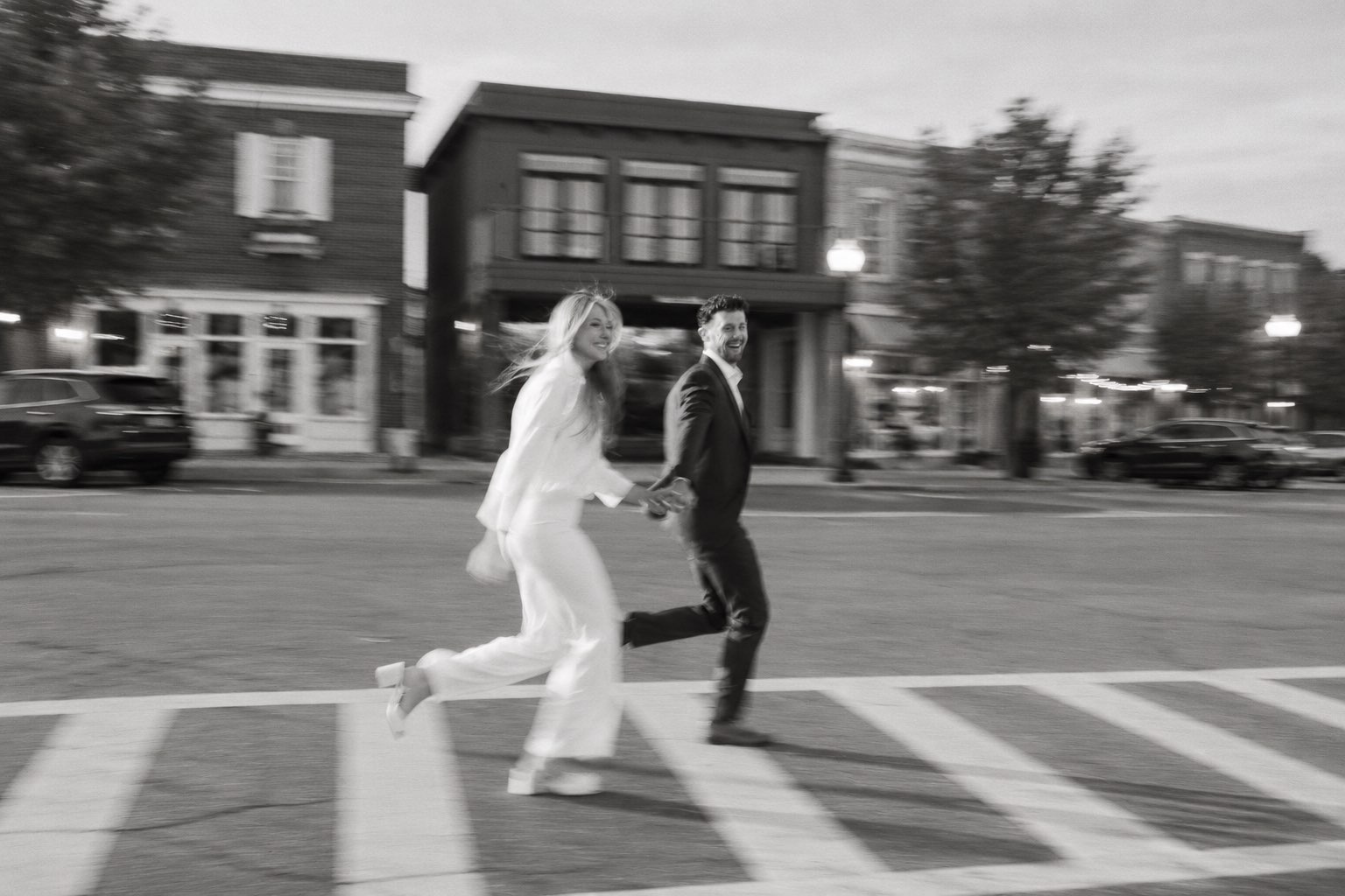 A couple running hand in hand across a street at dusk, laughing