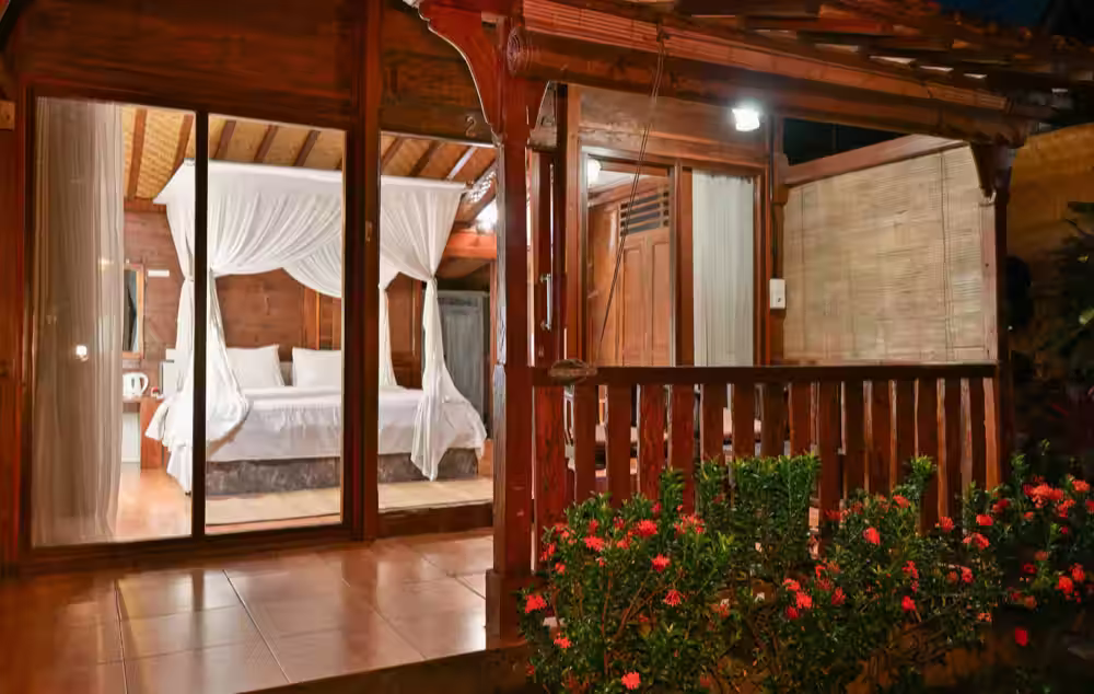 A timber villa at night, its glass doors open onto a four-poster bed hung with white netting